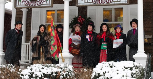 A group of carolers in winter attire sings on a snowy porch, surrounded by festive decorations.
