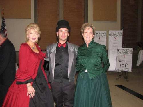 Three people in festive attire pose together at a holiday event, with banners for "Dickens Christmas Tale" in the background.