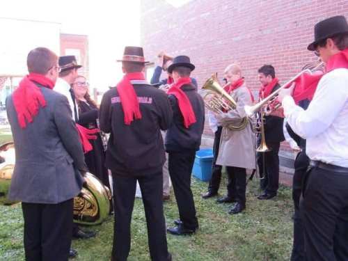 A group of musicians in hats and red scarves play instruments outdoors near a brick wall.