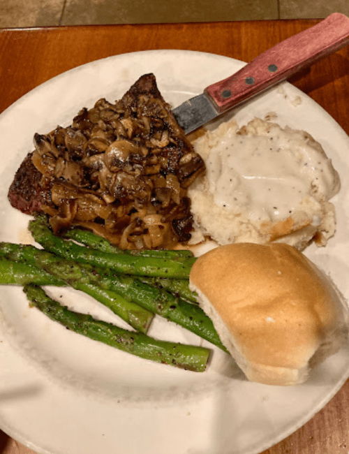 A plate with a steak topped with mushrooms, green asparagus, mashed potatoes with gravy, and a dinner roll.
