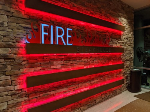 A modern restaurant sign reading "FIRE STEAKHOUSE" illuminated with red lights against a stone wall.