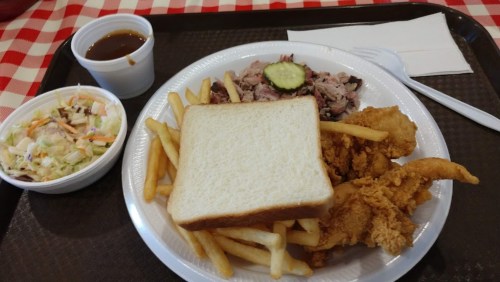 A plate with fried chicken, fries, coleslaw, a slice of bread, and a small cup of barbecue sauce on a checkered tablecloth.