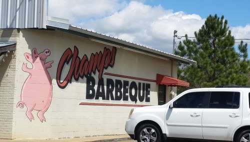 A restaurant building with a pig mural and the sign "Champs Barbeque" on the wall, with a white SUV parked nearby.