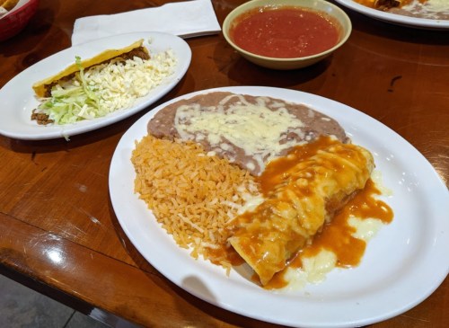 A plate of enchiladas with cheese sauce, rice, refried beans, and a taco on the side, with a bowl of salsa.