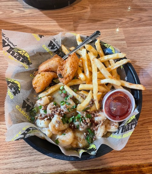 A plate of shrimp with toppings, fries, a fried item, and a small cup of dipping sauce on a wooden table.