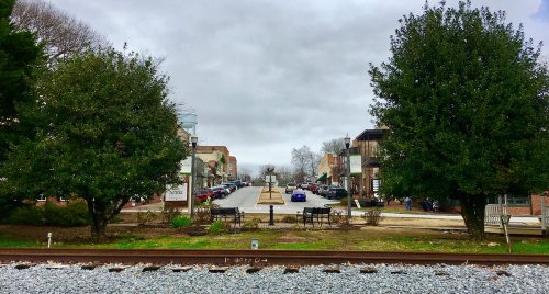 A view of a street lined with trees and shops, with a railway track in the foreground and cloudy skies above.