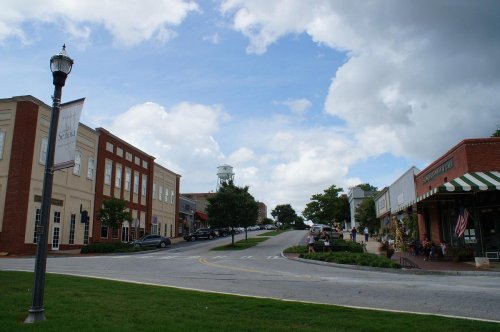 A charming small-town street with shops, greenery, and a water tower under a partly cloudy sky.