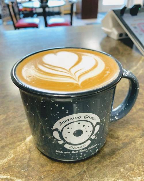 A close-up of a latte in a speckled mug with a heart-shaped foam design, placed on a wooden table.