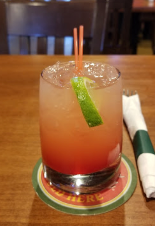 A colorful cocktail with a lime wedge and two straws, served in a glass on a wooden table.