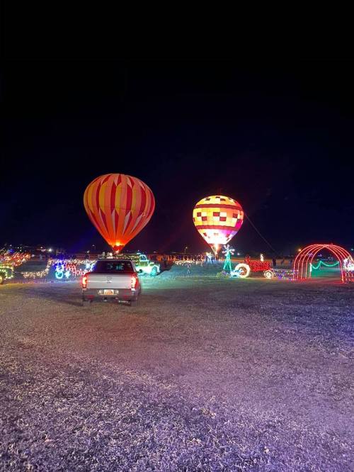 Colorful hot air balloons illuminated at night, surrounded by festive lights and a parked vehicle in the foreground.