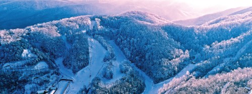Aerial view of snow-covered mountains and ski slopes under a soft pink and blue sky.