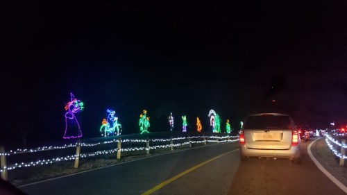 Colorful holiday lights depicting various festive figures along a dark road, with cars lined up in traffic.