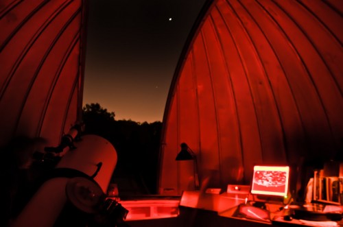A dimly lit observatory at night, featuring a telescope and a glowing computer screen, with stars visible outside.