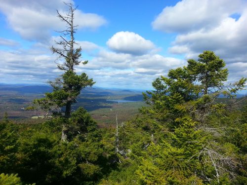 A scenic view from a mountain, featuring green trees, rolling hills, and a blue sky with fluffy clouds.