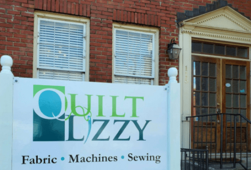Sign for "Quilt Lizzy" featuring fabric, machines, and sewing, in front of a brick building with windows and a door.