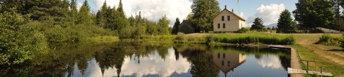 A serene landscape featuring a house by a pond, surrounded by trees and reflecting clouds in the water.