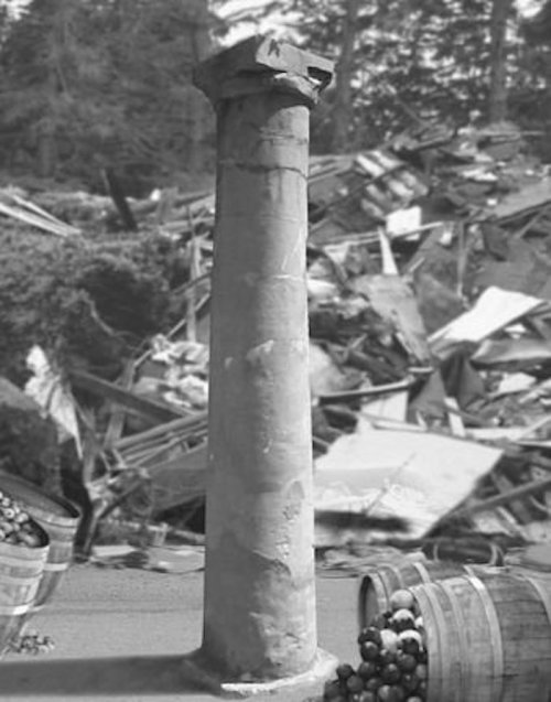 A weathered stone column stands in front of a chaotic background of debris and barrels of fruit.