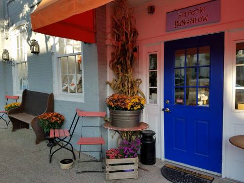 A charming storefront with a blue door, potted flowers, and rustic seating, adorned with autumn decorations.