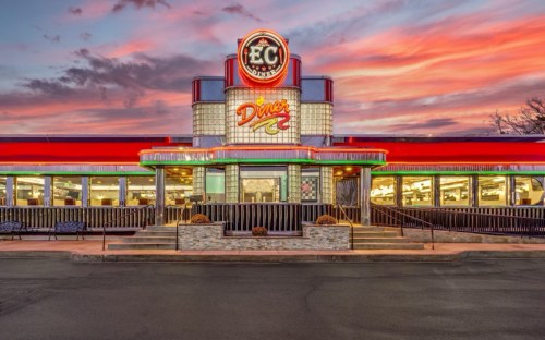 A vibrant diner with a retro design, illuminated by neon lights, against a colorful sunset sky.