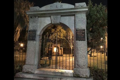 Stone archway with "DAR" inscription, featuring gates and plaques, set in a dimly lit park at night.
