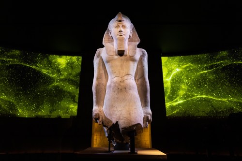 A large ancient statue of a pharaoh, illuminated against a vibrant green starry background.