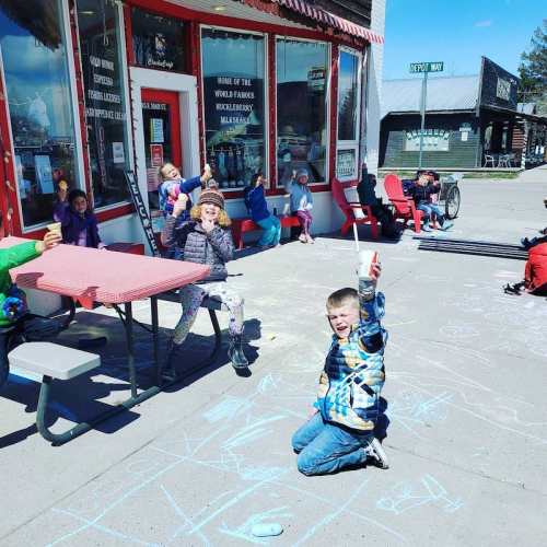 Children playing with chalk outside a shop, sitting at tables, enjoying a sunny day in a small town.
