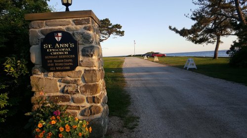 Sign for St. Ann's Episcopal Church beside a gravel path leading to the waterfront, with trees and a clear sky.