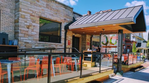 A modern café with a wooden deck, orange chairs, and large windows, set against a blue sky.
