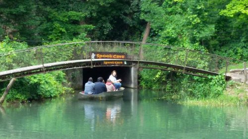 A group of people in a boat approaches a cave entrance under a bridge, surrounded by lush greenery.