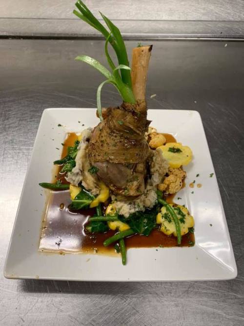A plated dish featuring a lamb shank on a bed of vegetables and creamy mash, garnished with green onions.