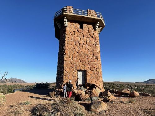 A stone tower stands against a clear blue sky, with people exploring its base in a desert landscape.