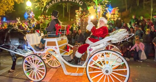 Santa Claus waves from a festive horse-drawn carriage, surrounded by holiday lights and a cheering crowd.