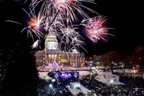 Fireworks illuminate the night sky over a snowy city square, with a large crowd gathered in front of a festive stage.