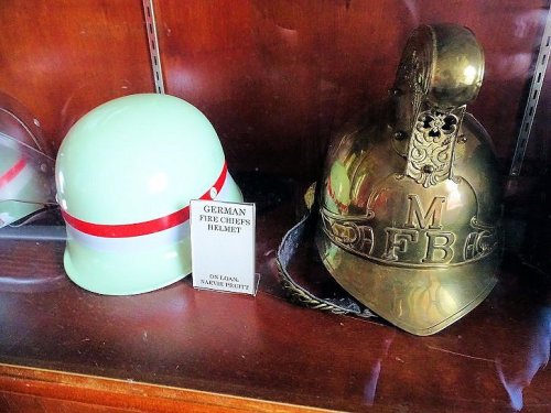 A display case featuring a German fire chief's helmet and a shiny brass helmet labeled "FB."