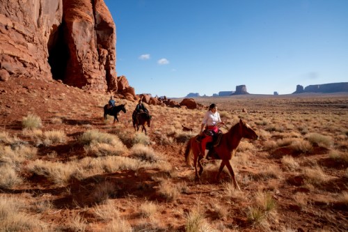 A group of horseback riders traverses a desert landscape with red rock formations and distant mesas under a clear blue sky.