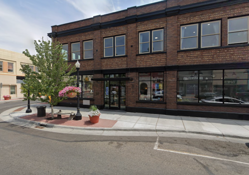 A brick building on a street corner with large windows, planters, and trees, featuring a sidewalk and street view.