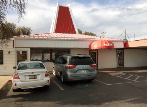 A fast-food restaurant with a red and white roof, featuring a large umbrella at the entrance and parked cars in front.