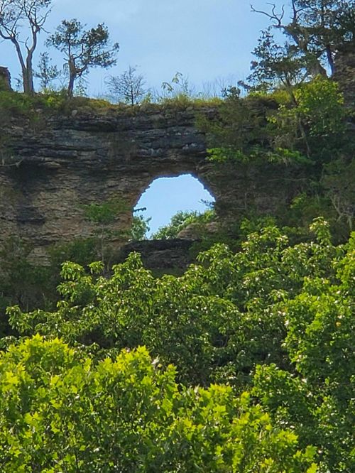A natural rock formation with a hole, surrounded by lush green trees and a clear blue sky in the background.
