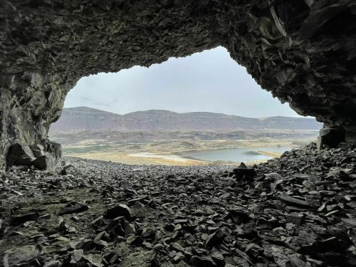 View from inside a rocky cave, overlooking a landscape of mountains and a body of water in the distance.