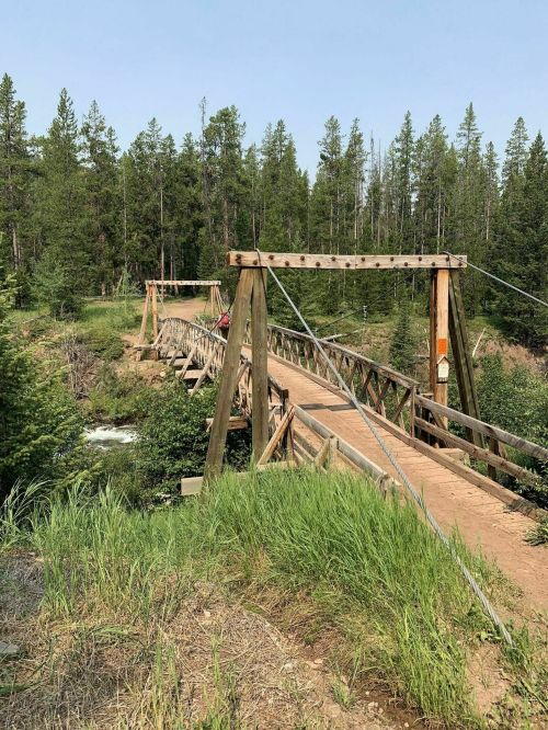 A wooden suspension bridge spans a river, surrounded by lush green trees and grass under a clear blue sky.