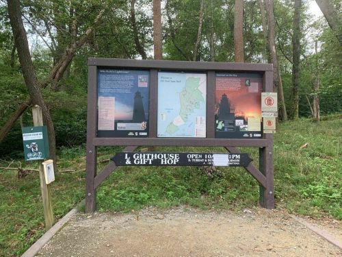 Information board at a lighthouse site, featuring maps and visitor details, surrounded by trees. Open hours displayed.