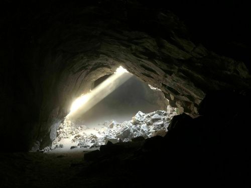 A dimly lit cave with a beam of sunlight illuminating the rocky interior and scattered stones on the ground.