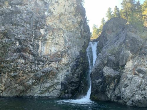 A waterfall cascading down rocky cliffs into a serene body of water, surrounded by trees and sunlight.