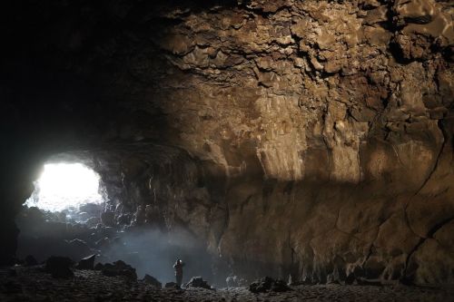 A person stands inside a large cave, illuminated by light streaming in from the entrance, highlighting the rocky textures.