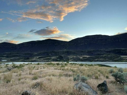 A serene landscape featuring a river, grassy terrain, and dark mountains under a colorful sky at dusk.