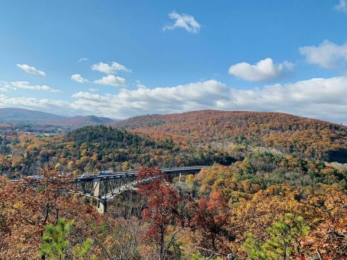 A scenic view of a bridge spanning a valley surrounded by vibrant autumn foliage and rolling hills under a blue sky.