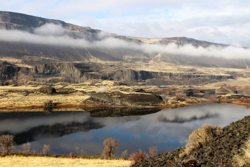 A serene landscape featuring a calm lake reflecting mountains and clouds, surrounded by grassy terrain and sparse trees.