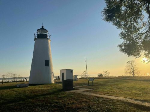 A white lighthouse stands on a grassy shore, with a clear sky and sun setting in the background.