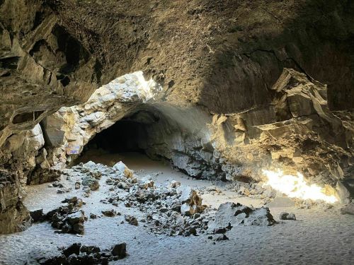 A dimly lit cave with rocky formations and a sandy floor, illuminated by shafts of light from above.