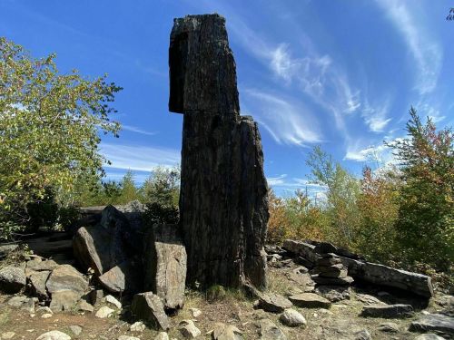 A tall, weathered rock formation stands amidst colorful trees under a blue sky with wispy clouds.